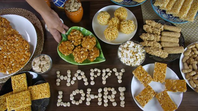 Hands of an Indian woman serving peanuts chikki on Lohri the harvest festival in India. Top view shot of 'Happy Lohri' text written with rewri surrounded by Lohri items like bhujia chikki  murmura ...