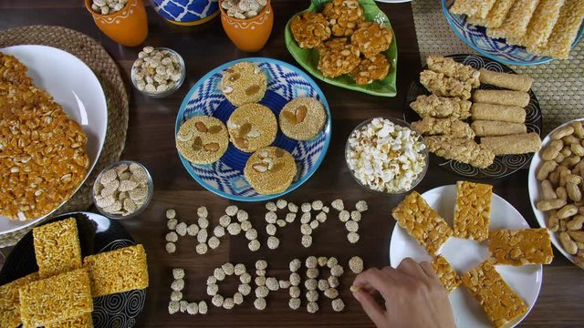Top view shot of beautifully plated edible items for the Lohri festival in India. Hands of an Indian woman writing 'Happy Lohri' with rewri on a wooden table surrounded by til patti  popcorns  gaja...