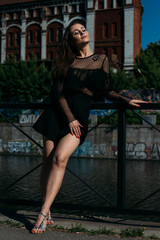 beautiful brunette stands on the bridge, near the river,in the city. a girl in a black dress puts a face to the sun and the wind that blows on her