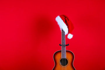 Fototapeta na wymiar Santa Clauses hat hanging on vintage style travel size acoustic guitar with rosewood neck and no pickguard over festive red wall background. Close up, copy space for text, top view.