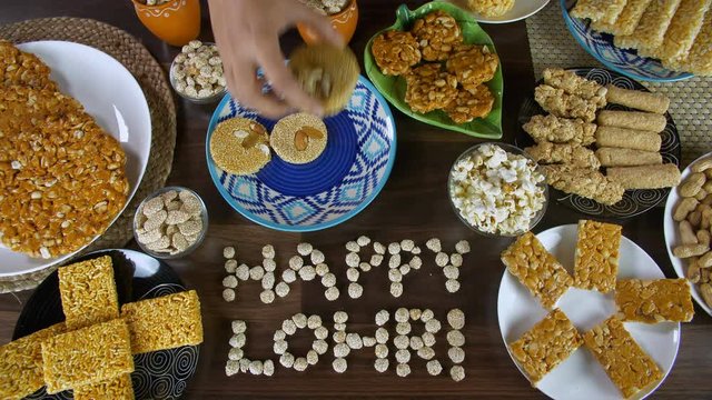 Woman hands serving til patti in a blue ceramic plate for the Lohri festival in India. Top view shot of 'Happy Lohri' text written with rewri and surrounded by Lohri items like gajak  peanuts chikk...