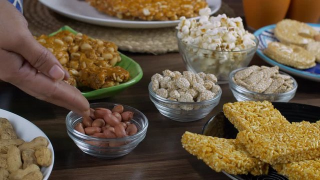 Beautifully served Lohri items consumed on the Lohri festival during wintertime. Closeup shot of Indian woman hands pouring peanuts in a glass bowl surrounded by rewri  popcorns  peanuts chikki  bh...
