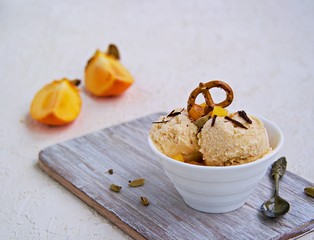 Dessert, homemade ice cream or frozen yogurt with persimmons, decorated with salted pretzels and grated chocolate, in a white bowl on a light concrete background. Selective focus.