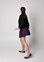full length portrait of a pretty brunette girl wearing a school uniform of black jacket and plaid skirt. Standing pose, with back to the camera, isolated against a studio background.