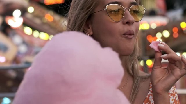 A positive cute woman is eating candyfloss while walking outdoors in amusement park