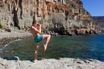 Fighter practicing knee kick and arms defense outdoors. Young sports man training by the sea with rocky mountain on background on sunny day in Puerto Mogan, Gran Canaria