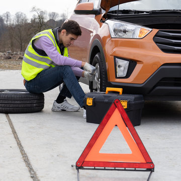 Caucasian young man wearing a high-visibility or hi-vis vest and jeans changing a tire sitting on stepney near car on the roadside. Male changes a damaged wheel on the rural road.