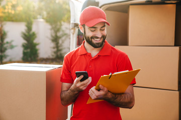 Image of happy delivery man holding clipboard and using cellphone