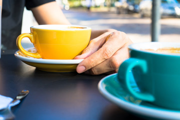 A yellow cup of coffee on the table. Young woman drinks fresh morning coffee in downtown street cafe.  