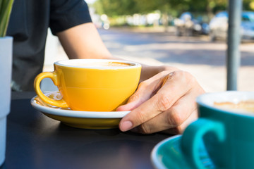 A yellow cup of coffee on the table. Young woman drinks fresh morning coffee in downtown street cafe.  