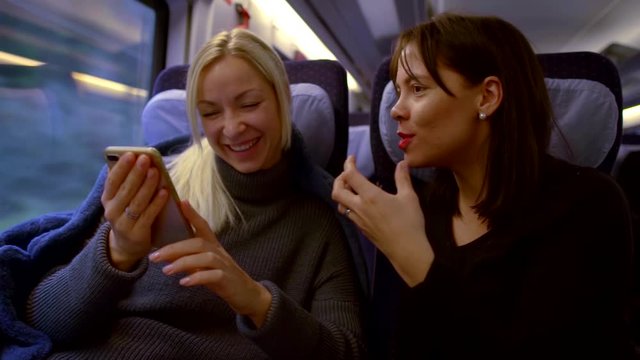two women are on the train. a brunette with bright lipstick on her lips is saying something. the blonde looks at her smartphone. girlfriends have fun laughing