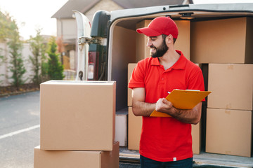 Image of smiling delivery man writing while standing with parcel boxes