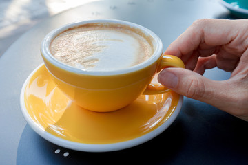 A yellow cup of coffee on the table. Young woman drinks fresh morning coffee in downtown street cafe.  