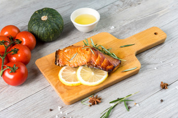 Grilled salmon steak on vintage wooden board with fork, olive oil, vegetables, herbs and spices on dark concrete background. Raw salmon fillet with vegetables, spices and lemon on wooden table.