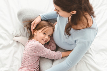 Little girl hugging pregnant mom's belly while lying on bed