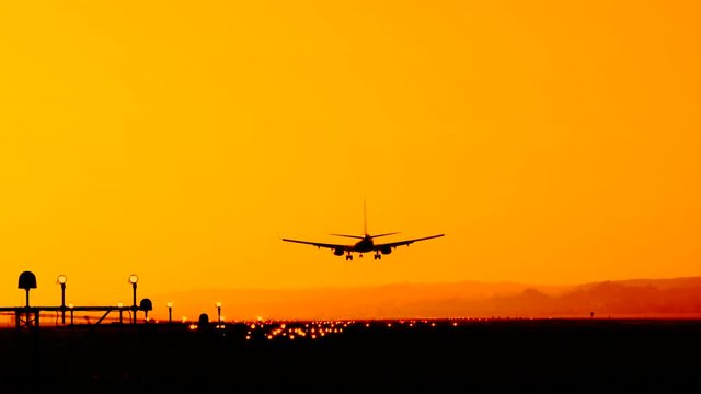 Back view silhouette of airplane landing at the airport at sunny orange and purple sunset. Runway end identification lights and strobe glowing.