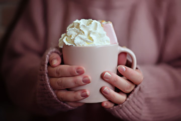 Dusty pink mug with coffee with cream and marshmallows in the hands of a woman in a warm pink knitted sweater.