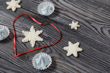 Marshmallows and snowflakes from marshmallows. Nearby is a scarlet ribbon laid in the shape of a heart. On old pine boards painted black.