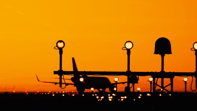 Silhouette of airplane at the airport at sunny orange and purple sunset. Plane taxiing on the runway preparing for take off. Runway end identification lights and strobe glowing.
