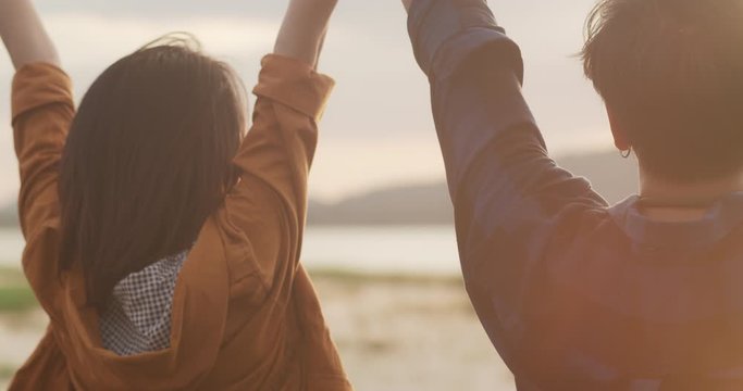 Young asia campers couple standing raised arms at campsite near beach. Happy male and female traveler success and fun when sunset in evening. Outdoor activity, adventure travel, or holiday vacation.