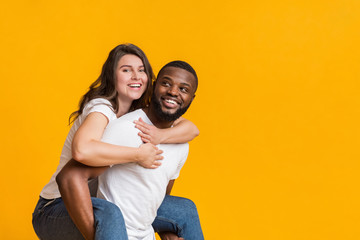 Cheerful black guy piggibacking his girlfriend, interracial couple having fun together