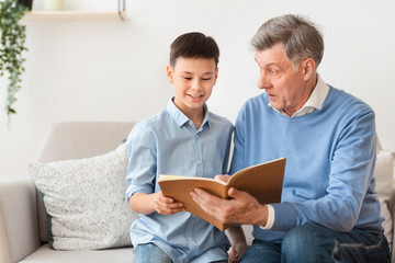 Grandfather And Grandson Reading Book Sitting On Sofa At Home