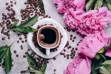 Coffee in a white cup around coffee beans and flowers peonies of pink color with green leaves. Top view