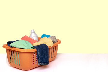 Closeup of a orange wash basket with colorful dirty clothes and a white detergent bottle on a bright table against light yellow background. Space for product montage.