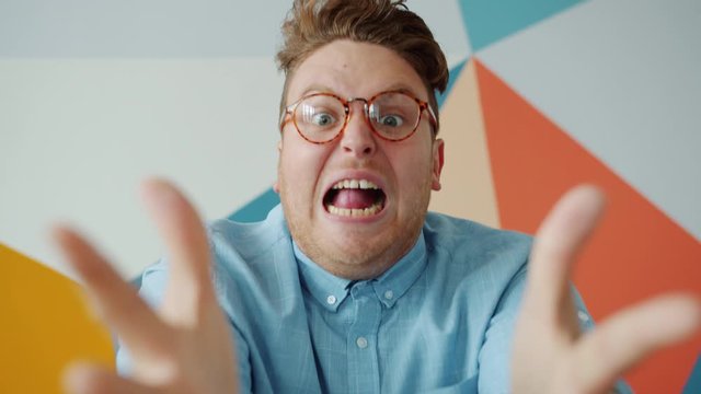 Angry guy in glasses is yelling holding head gesturing looking at camera on colorful background. Negative emotions, feelings and modern people concept.
