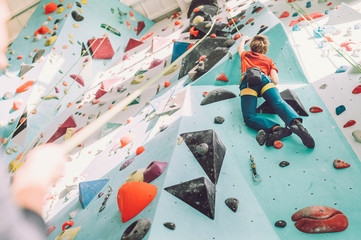 Teenager boy at indoor climbing wall hall. Boy is climbing using a top rope and climbing harness and somebody belaying him from floor. Active teenager time spending concept image. - obrazy, fototapety, plakaty