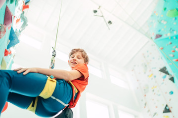 Teenager boy at indoor climbing wall hall. Boy is climbing using a top rope and climbing harness and somebody belaying him from floor. Active teenager time spending concept image.