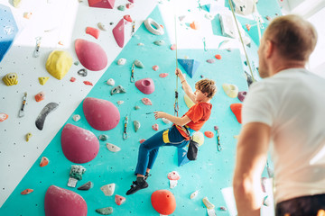 Father and teenager son at indoor climbing wall hall. Boy is hanging on the rope using a climbing harness and daddy belaying him on the floor using a belay device. Happy parenting concept image. - obrazy, fototapety, plakaty