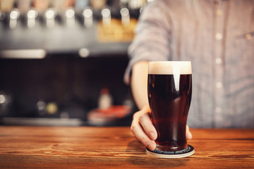 Bartender holds out glass of dark beer client behind wooden bar counter