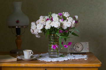 Bouquet with wild rose and blooming peas in a transparent vase on an old table. Near the old clock and lamp of the times of the Soviet Union. On the table is an open book and a white cup with coffee.