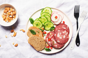 Appetizer with fresh vegetables cucumber, radishes, spinach, micro-greens, and slices of salami on a dark plate in hard bright light. Trend snapshot. Top view. Hard shadows
