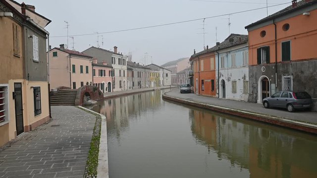 Comacchio, Italy. December 2019. Static shooting on a pretty canal: the houses are reflected in the water. The canal curves hiding between the houses and the fog. Nobody.
