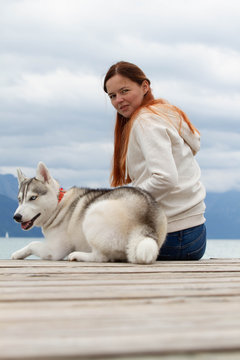 A young woman with brown hair and white sweater is sitting at the pier at the lake with calm water. A Siberian husky female dog is lying down near the girl. The Alps mountains in the background.