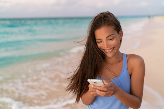 Asian Woman Using Smart Phone Texting Online On Dating App Holding Cellphone Mobile Phone Outside Walking On Beach At Sunset. Happy Young Eurasian Girl.