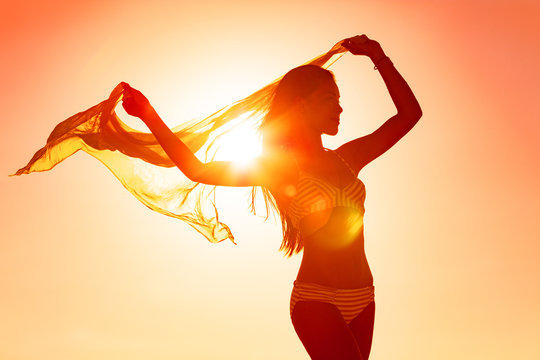 Wellness woman body silhouette curves dancing carefree with femininity with scarf flowing in the wind at sunset glow flare for life happiness. Sun vacation travel beach.