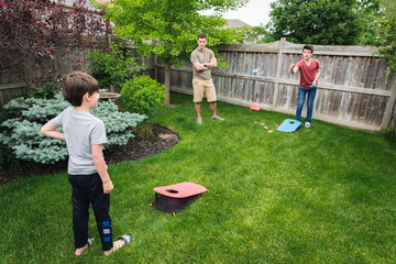 Two boys playing corn hole game in backyard with dad watching them.