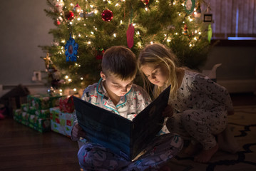 Boy reading a book to girl in front of Christmas tree