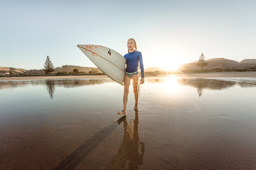 Preteen girl holding surfboard on beach at sunset