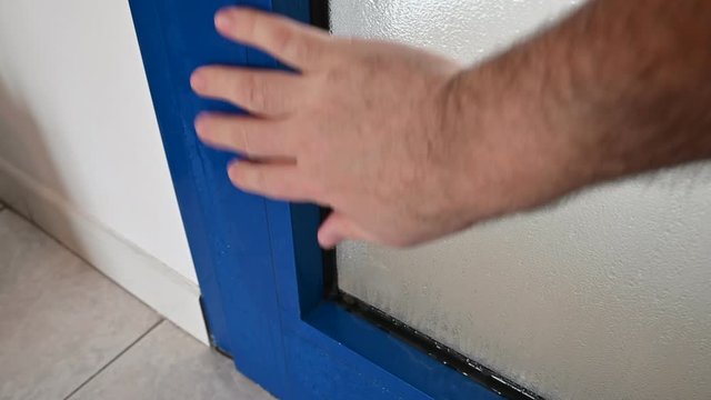 Thermal insulation problems cause condensation on the door window frame: the Caucasian man's hand passes over the blue metal frame, highlighting the formation of humidity.