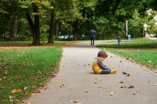 A small boy sits on a leaf-strewn path as his family walks on