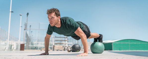 Fitness man banner panorama. Athlete strength training pushup balancing legs on medicine ball for advanced core body workout push-ups floor exercises at outdoor gym.