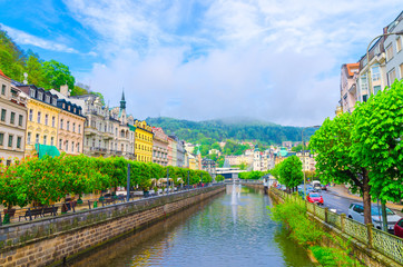 Fototapeta na wymiar Karlovy Vary (Carlsbad) historical city centre with Tepla river central embankment, colorful beautiful buildings, Slavkov Forest hills with green trees background, West Bohemia, Czech Republic