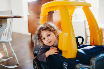 Young girl looking at camera sitting in toy car inside dining room