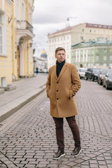 Attractive blond caucasian man in beige coat, with dark blue scarf tied around his neck, brown pants and dark leather shoes keeps hands in pockets. Selective focus on pretty hipster. Lifestyle concept