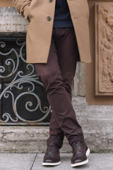 Close up photo of unrecognizable pretty male in fashionable wool autumn beige coat, brown pants and dark leather shoes with white soles. Young stylish man is standing against wall. Lifestyle concept.