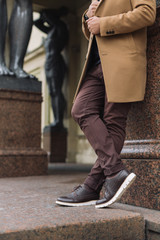 Cropped portrait of unrecognizable handsome man in elegant autumn beige coat, brown pants and dark leather shoes with white soles. Young stylish hipster is leaning against the wall. Fashion concept.
